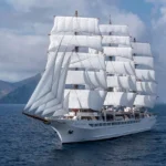Historic four-masted sailing yacht Sea Cloud under full sail at sea, showcasing its classic rigging and white sails against a coastal backdrop