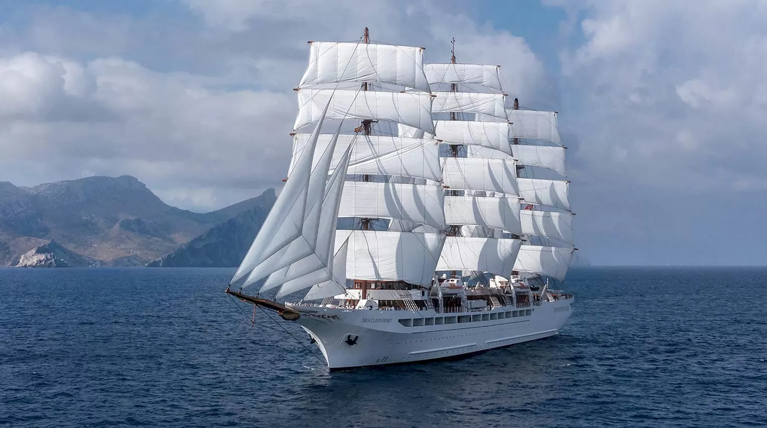 Historic four-masted sailing yacht Sea Cloud under full sail at sea, showcasing its classic rigging and white sails against a coastal backdrop