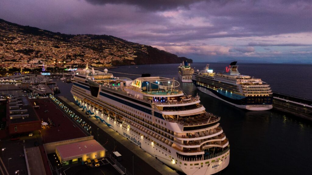 Multiple cruise ships docked at Funchal harbor at dusk, with illuminated vessels lined along the Madeira waterfront and city lights glowing in the background.