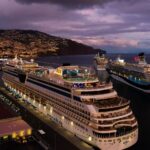 Multiple cruise ships docked at Funchal harbor at dusk, with illuminated vessels lined along the Madeira waterfront and city lights glowing in the background.
