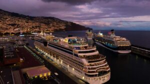Multiple cruise ships docked at Funchal harbor at dusk, with illuminated vessels lined along the Madeira waterfront and city lights glowing in the background.