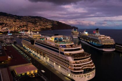 Multiple cruise ships docked at Funchal harbor at dusk, with illuminated vessels lined along the Madeira waterfront and city lights glowing in the background.