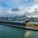 Aerial view of Hong Kong’s Kai Tak Cruise Terminal with the city skyline and Victoria Harbour in the background, highlighting the waterfront cruise facility and surrounding urban landscape.